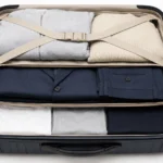 Hands folding a navy suit jacket by turning one shoulder inside out on a light wooden table to prepare it for packing.