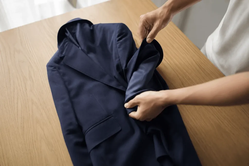 How to Pack a Suit in a Suitcase Without Wrinkles 1 Suit jacket wrapped around a rolled grey sweater with suit trousers partially folded around it inside a beige suitcase.