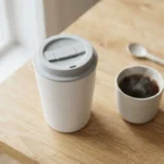 White ceramic travel mug with a slide-lock lid next to a steaming cup of coffee on a wooden desk
