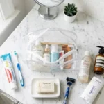 Flat lay showing what includes toiletries, with toothpaste, shampoo, soap, deodorant, razor, and lotion beside an open clear toiletry bag